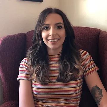 A smiling woman with long brown hair wearing a striped t-shirt sits in a red chair, showing a tattoo on her left arm.