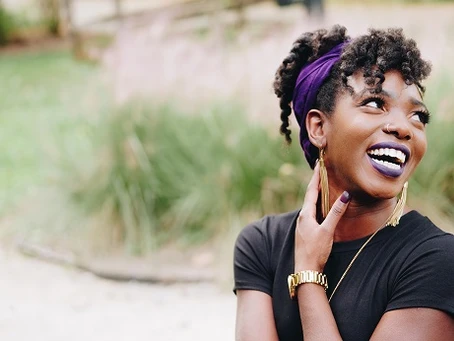 A joyful woman with curly hair tied up using a purple scarf, wearing a black top and a gold watch, smiling broadly and looking upwards while touching her cheek, against a blurred natural background.