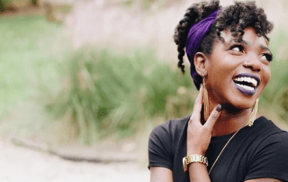 A joyful woman with a purple headband and dark curly hair styled in a bun, wearing a black top, laughing and looking up, her hand touching her neck, outdoors with greenery in the background.