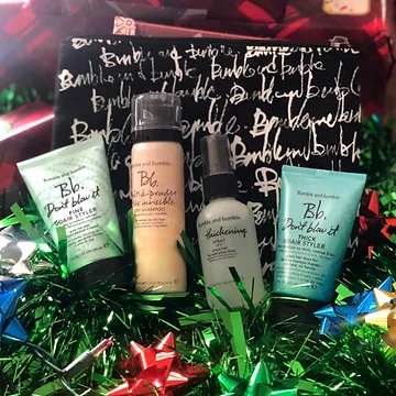 Four bumble and bumble hair products displayed on colorful tinsel, with a branded black and white gift box in the background, set in a festive, holiday-themed arrangement.