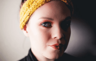 Close-up portrait of a woman with a yellow headband, red eye makeup, and neutral expression, focusing intently towards the camera against a blurred background.