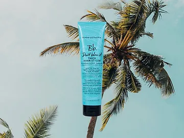 A tube of bumble and bumble styling cream held up against a clear sky, with palm trees in the background, evoking a tropical mood.