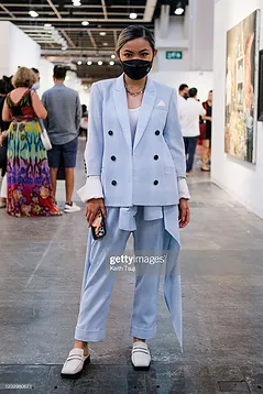 A stylish woman wearing a light blue double-breasted suit and mask stands in an art gallery, holding a phone and bag. she is surrounded by art viewers and paintings.