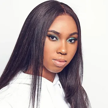 A woman with straight long black hair and striking green eyeshadow looks directly at the camera, wearing a white shirt against a plain light background.