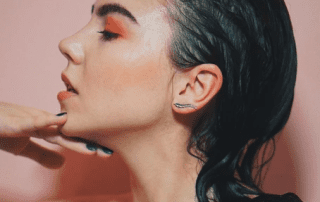 Close-up side view of a woman with slicked-back wet hair, wearing orange eyeshadow and silver highlighter. her hand gently touches her jawline, showcasing blue nail polish. background is soft pink.