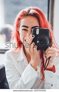 A woman with vibrant red hair and a white blazer enthusiastically taking a photo with a black digital camera, partially covering her face, near a window.