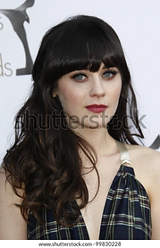 A woman with long brown hair featuring bangs, wearing a halter neck top with a plaid pattern, posing at a red carpet event, with soft-focus background.