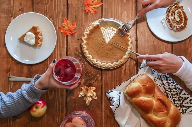 Top-down view of a wooden table with two people enjoying an autumn feast, featuring pumpkin pie, bread, a cranberry drink, and other seasonal treats surrounded by fall leaves.