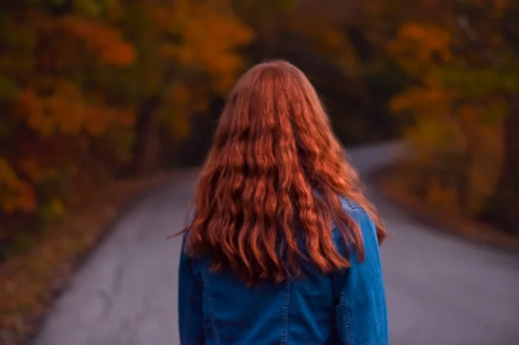 A woman with long red hair and a blue denim jacket standing on a road, surrounded by trees with vibrant autumn leaves. the focus is on her hair and the background is softly blurred.