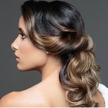 Profile view of a woman with an elegant braided updo hairstyle, featuring cascading brown and blonde highlights against a gray background.