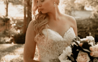 A bride in a lace wedding gown holds a bouquet of cream and blush roses, bathed in soft sunlight filtering through trees.