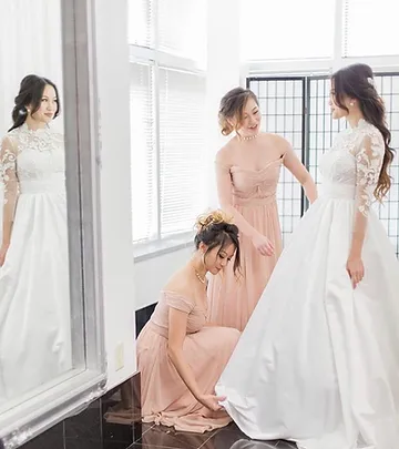 Three women in elegant dresses inside a bright room; one adjusts another's long white wedding gown while a third observes, all near large windows.