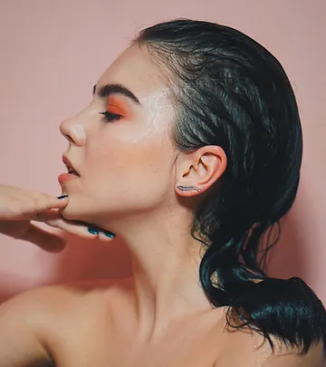 A woman with a side profile, showcasing a colorful makeup look with orange eyeshadow and highlighter, against a pink background. she has dark hair styled in a wet look and poses elegantly.