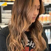 A woman with long wavy highlighted hair looks downward, partially revealing a graphic t-shirt, in an indoor setting with blurred background.