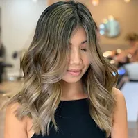 A woman with shoulder-length, wavy, balayage hairstyle in shades of brown and blonde, looking downward with a subtle smile, indoors.