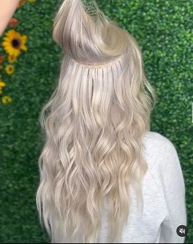 A woman showcases her long, ash blonde hair styled in soft waves, with one section artistically twisted and clipped at the back, against a background of sunflowers.