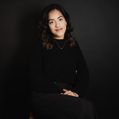 A woman with shoulder-length curly hair, wearing a black sweater and pants, sits on a stool against a dark background, smiling subtly at the camera.