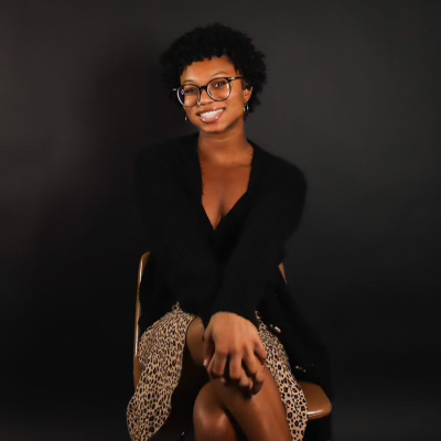 A woman with short curly hair and glasses smiles cheerfully, sitting relaxed in a chair. she's wearing a black cardigan and a leopard print skirt, against a dark background.