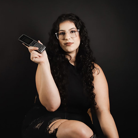 A woman with curly hair and glasses, wearing black, holds an old flip phone while sitting and looking at the camera against a dark background.