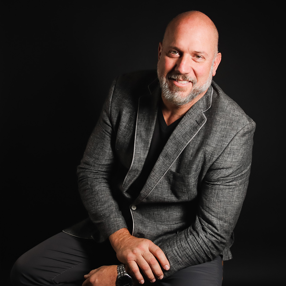 A bald man with a beard, wearing a gray blazer, smiling and sitting with his arms crossed against a black background.