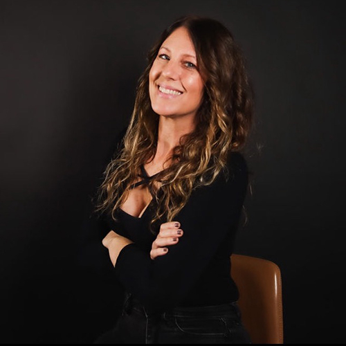 A smiling woman with long, curly hair sits with crossed arms in a black top, against a dark background.