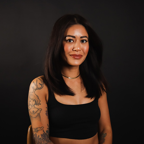 A woman with shoulder-length dark hair and tattoos on her arms and shoulder sits against a black background, wearing a black tank top and a choker necklace, looking at the camera.