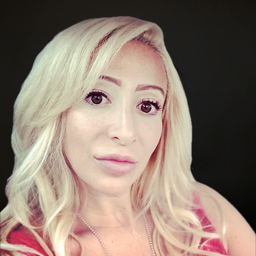 A portrait of a woman with long blonde hair, wearing subtle makeup and a necklace, set against a dark background. she has a calm expression.